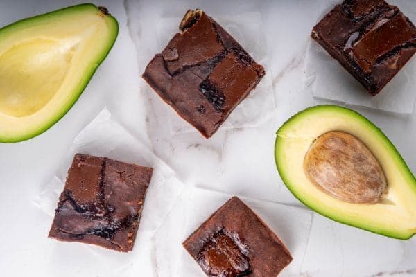 Four high protein avocado brownies, surrounded by two halves of an avocado.