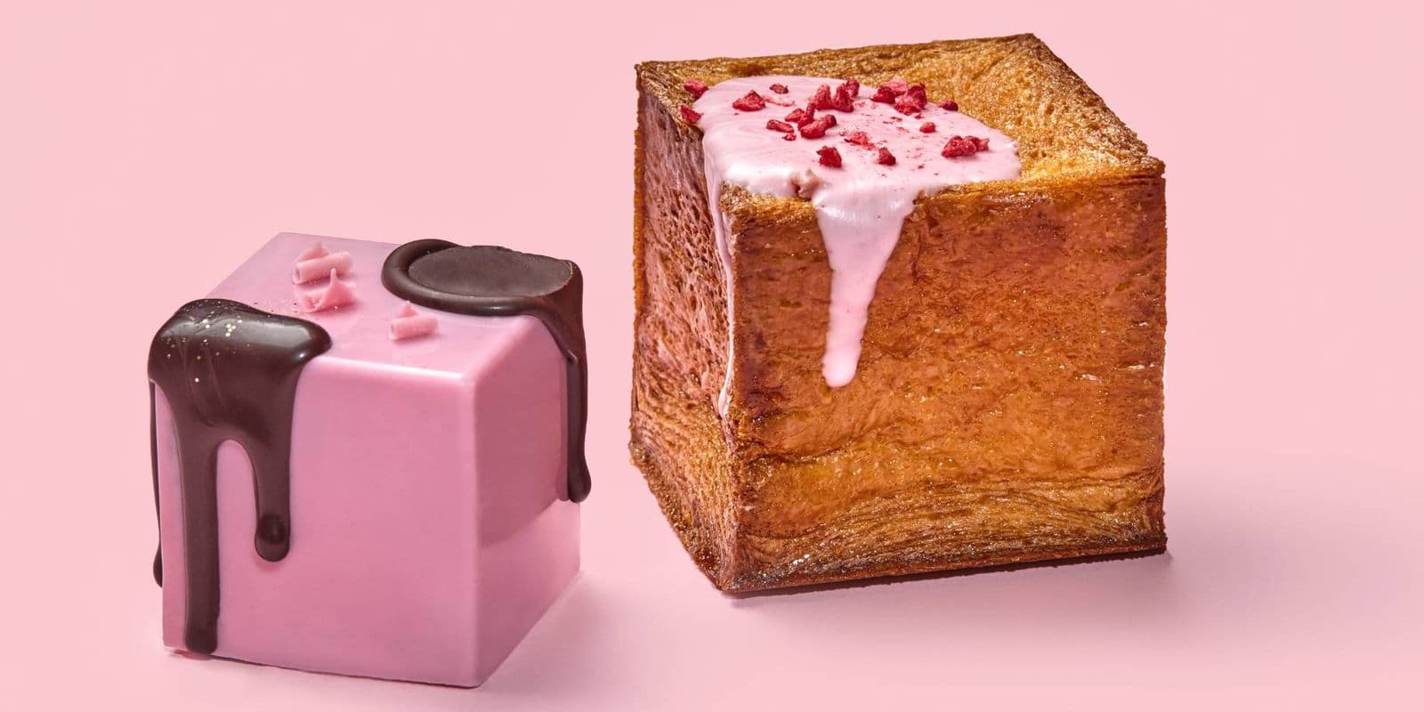 Two cubed crossiants, one covered in a pink coating and drizzled with chocolate, the other plain with a light pink drizzle and dried raspberry pieces.