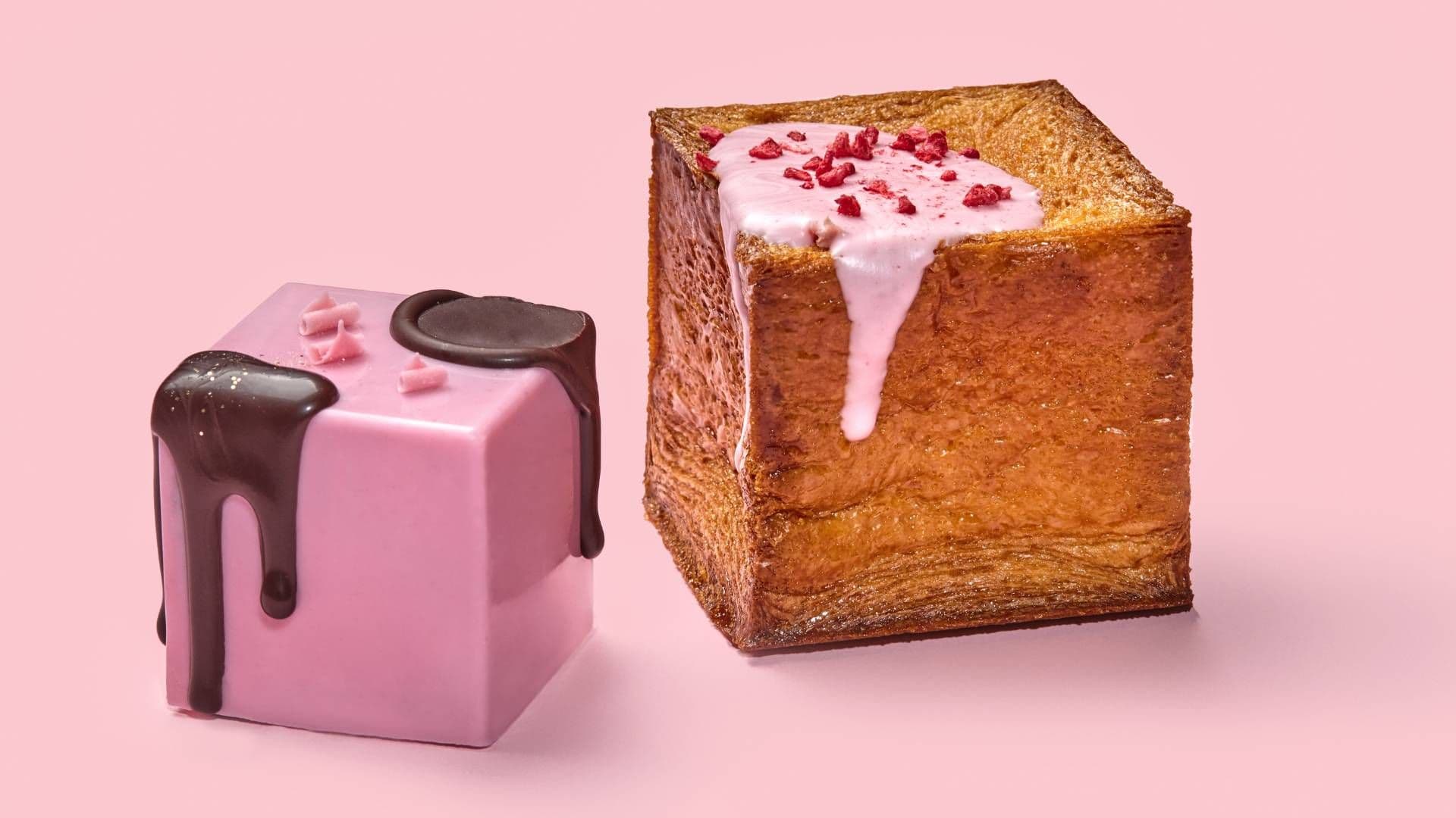 Two cubed crossiants, one covered in a pink coating and drizzled with chocolate, the other plain with a light pink drizzle and dried raspberry pieces.