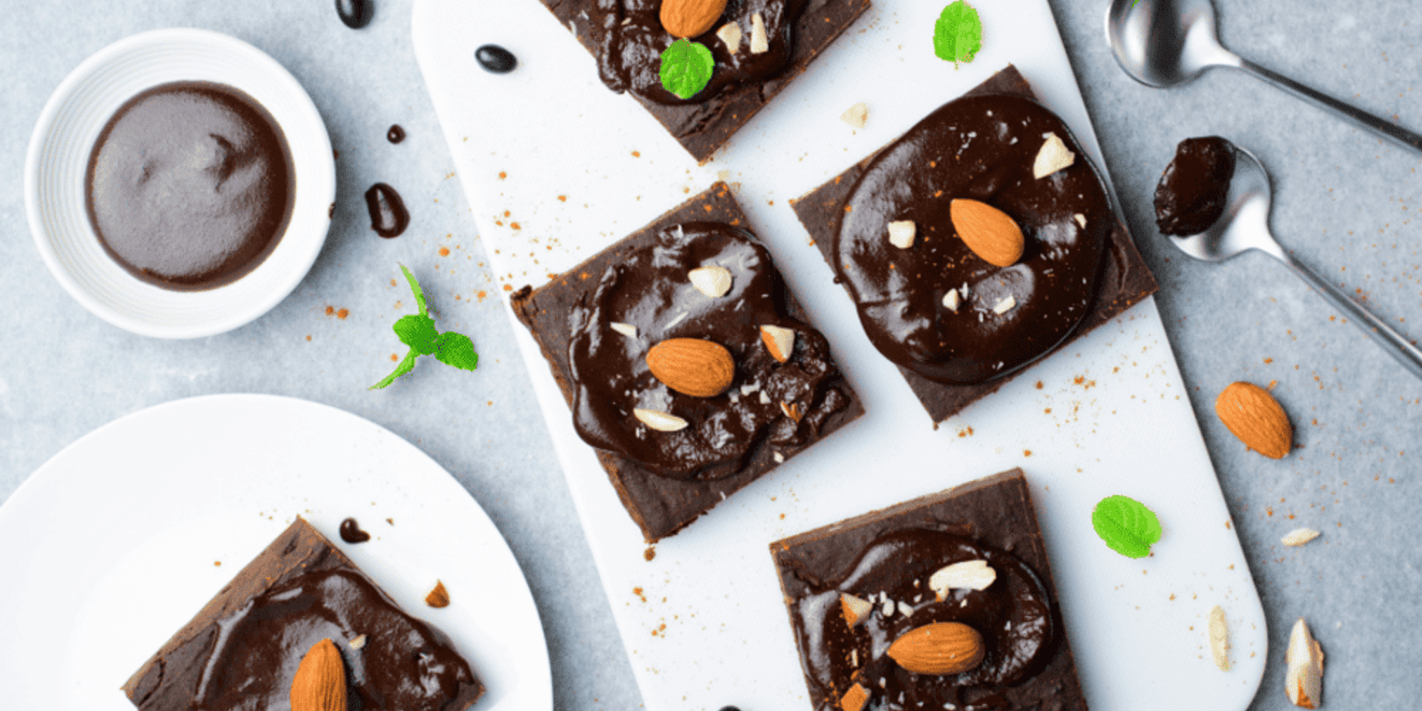 Five healthy, sugar-free brownies topped with chocolate sauce and almonds on a white chopping board.