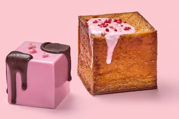 Two cubed crossiants, one covered in a pink coating and drizzled with chocolate, the other plain with a light pink drizzle and dried raspberry pieces.