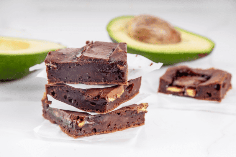 Three vegan chocolate brownies stacked on each other, with an open avocado half in the background.
